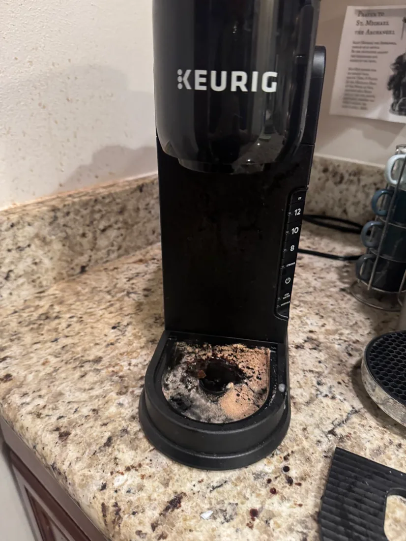 A coffee-stained single-serve coffee maker with used grounds in the drip tray before cleaning